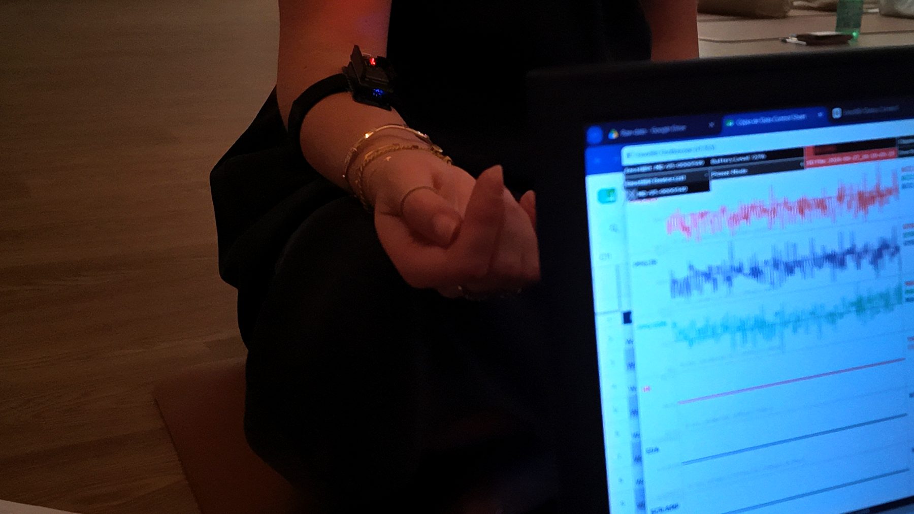 A participant's wrist with a biometric sensor, next to a laptop showing live heart and skin-response data