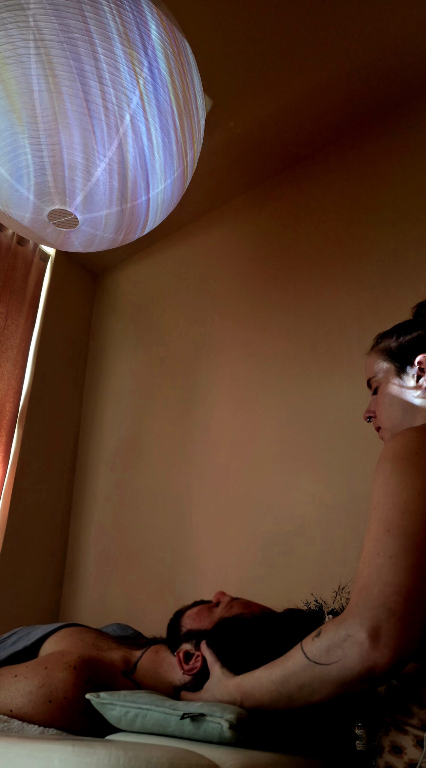 A practitioner's hands support a client's head during a one-on-one session, under a paper lantern glowing with projected visuals